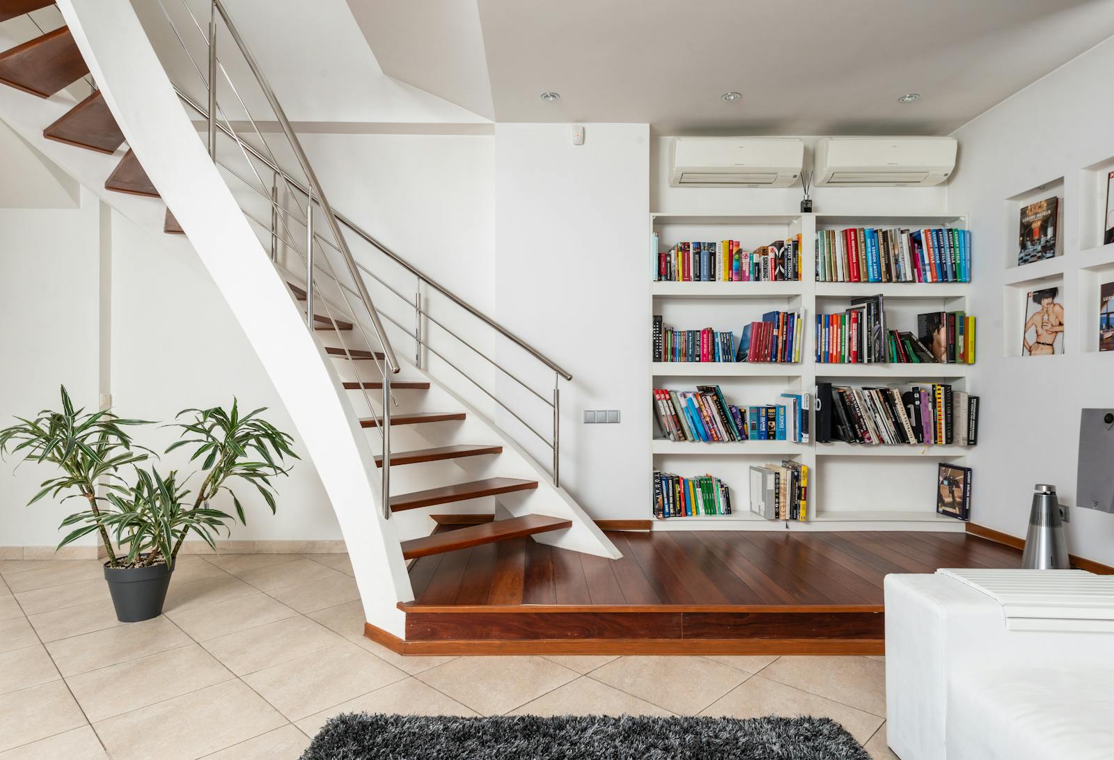 Assorted textbooks on shelves against contemporary staircase above ceramic floor with potted plant and carpet at home